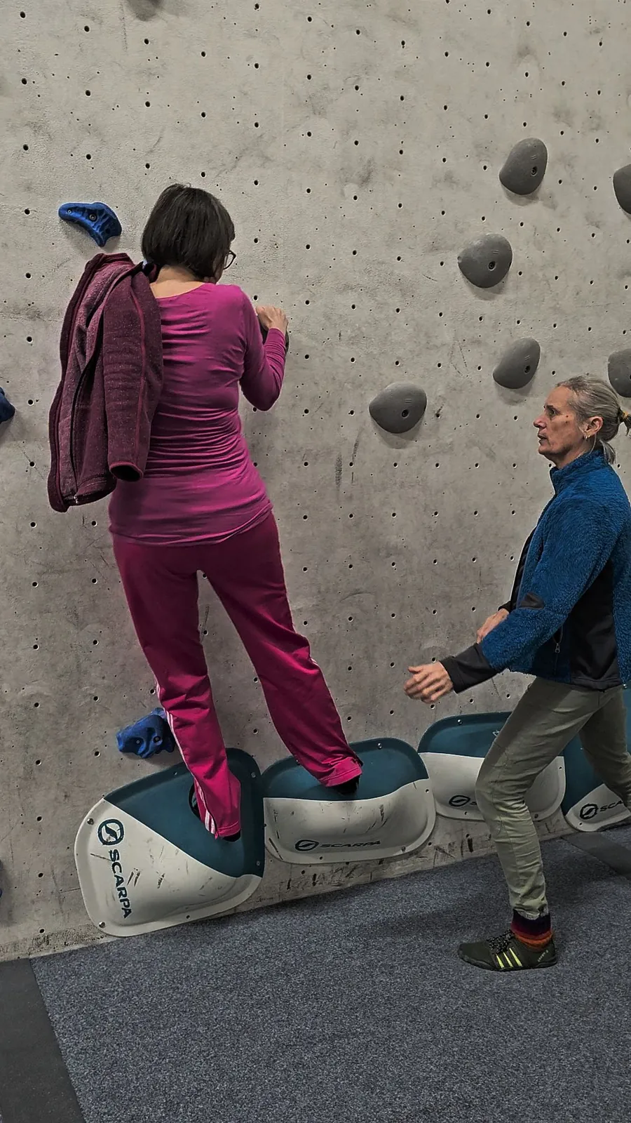 Eine Teilnehmerin steht auf großen Tritten knapp über dem Boden an der Boulderwand. Helga steht dicht hinter ihr und sichert mit den Händen.