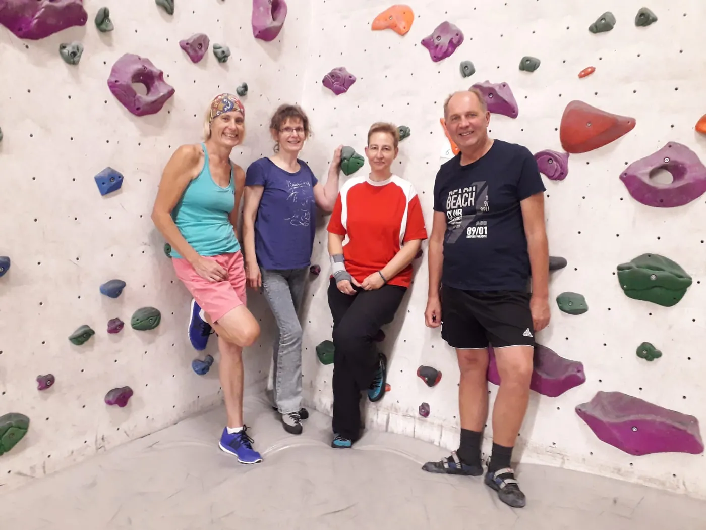 Vier Menschen stehen lachend vor einer bunten Boulderwand, ganz links Helga mit Stirnband.